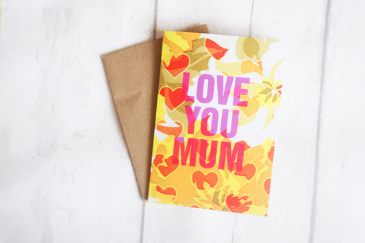 Greeting card with 'Love You Mum' text on a yellow background with hearts, next to a brown envelope on a white surface.