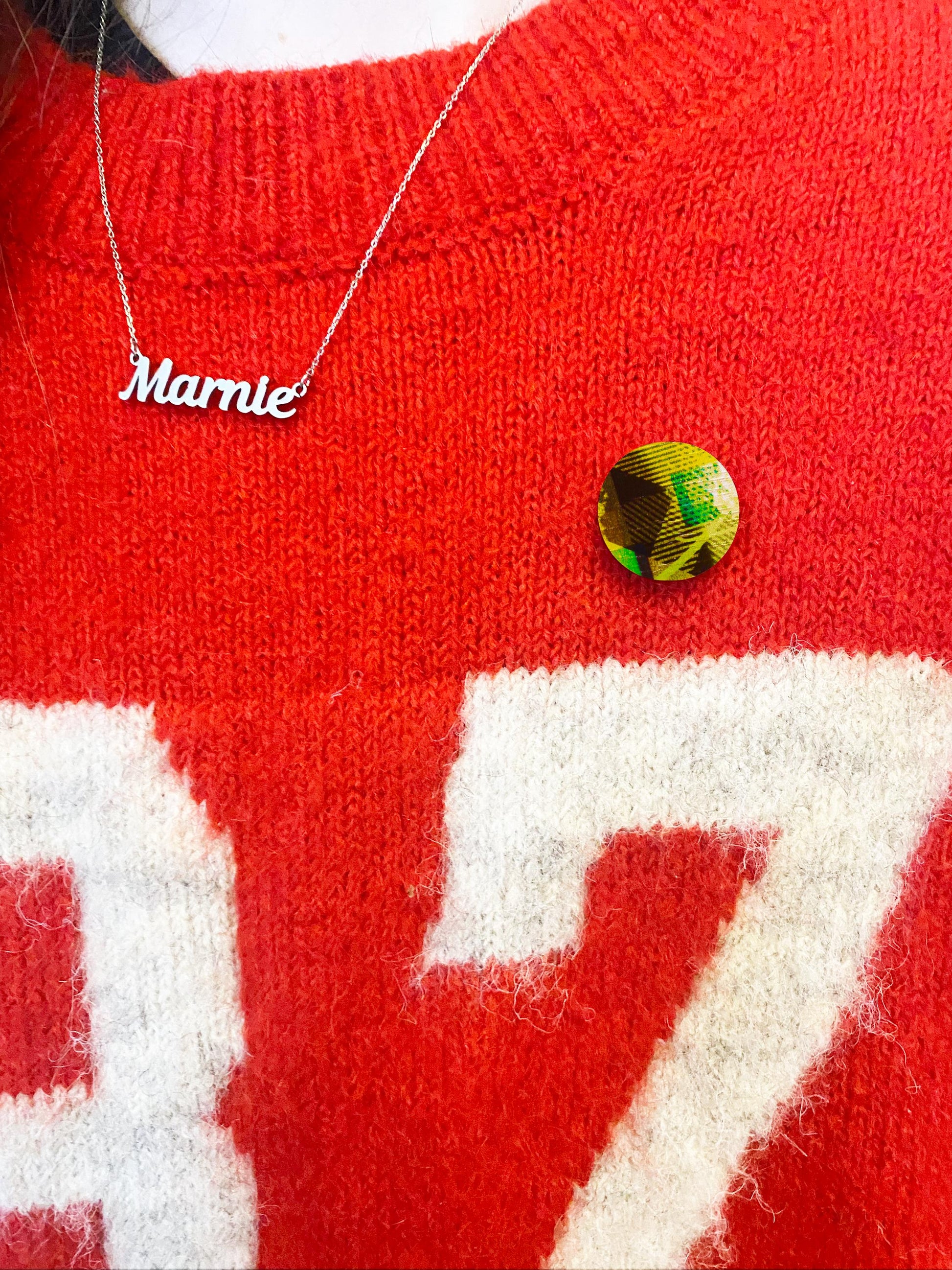 Red sweater with white text and a green pin badge, featuring a necklace with 'Marnie' on it.