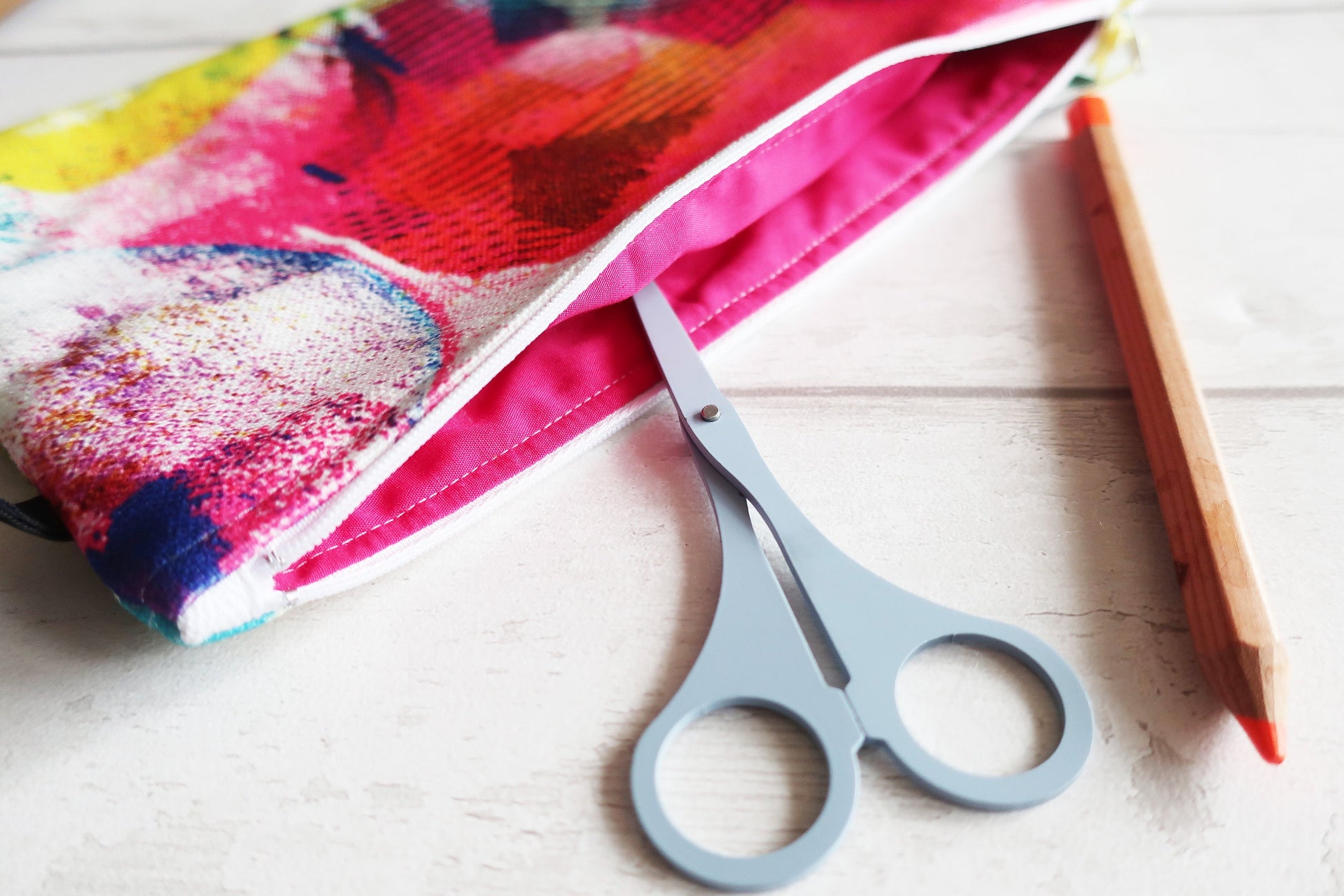 Pair of scissors on a white surface with a colorful fabric and pencil in the background