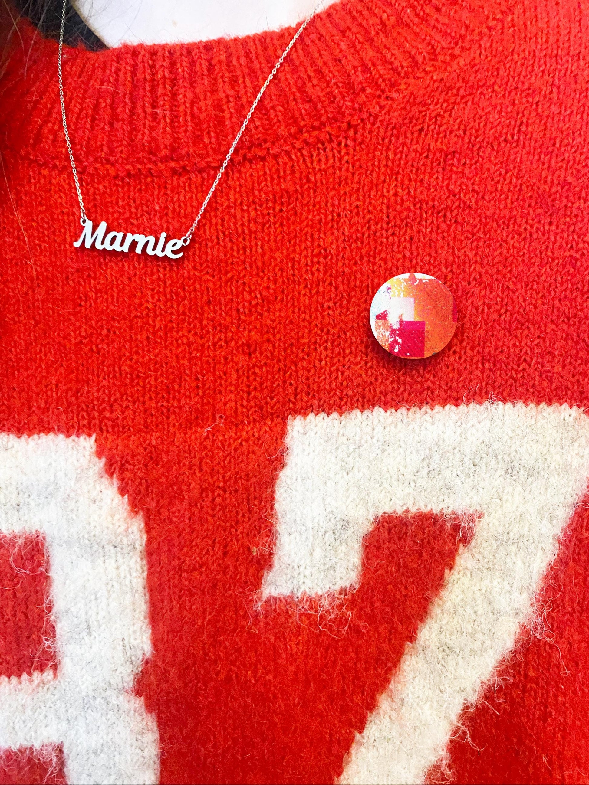 Red sweater with 'Marnie' necklace and red pin badge, close-up view.
