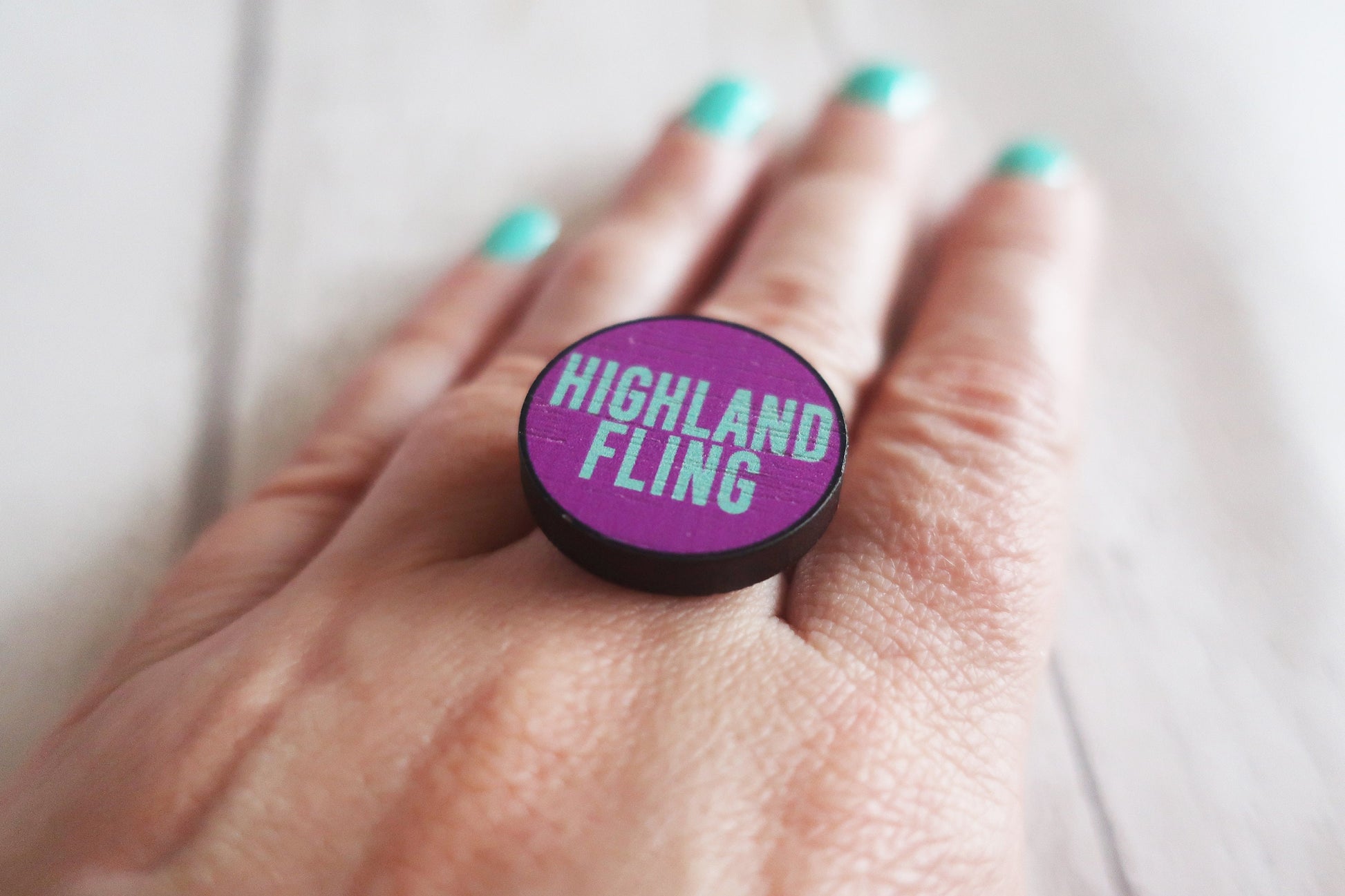Purple ring with 'Highland Fling' text held in a hand with teal nail polish.