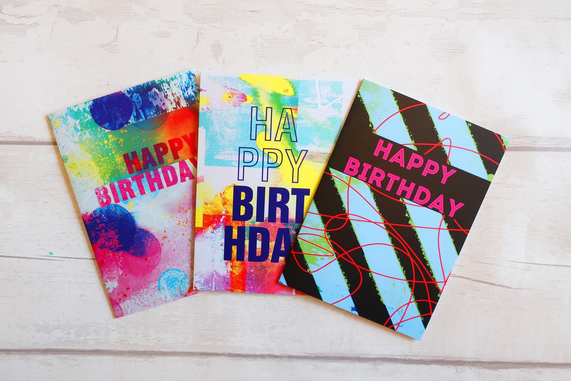 Three colorful birthday cards with abstract designs on a light wooden surface.