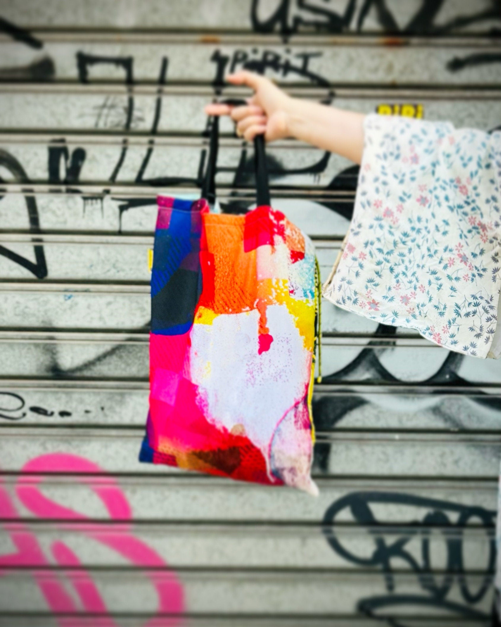 Colorful abstract tote bag held by a person in front of a graffitied wall.