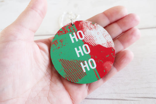 Hand holding a round, red and green necklace with 'HO HO HO' text on a light wooden surface.