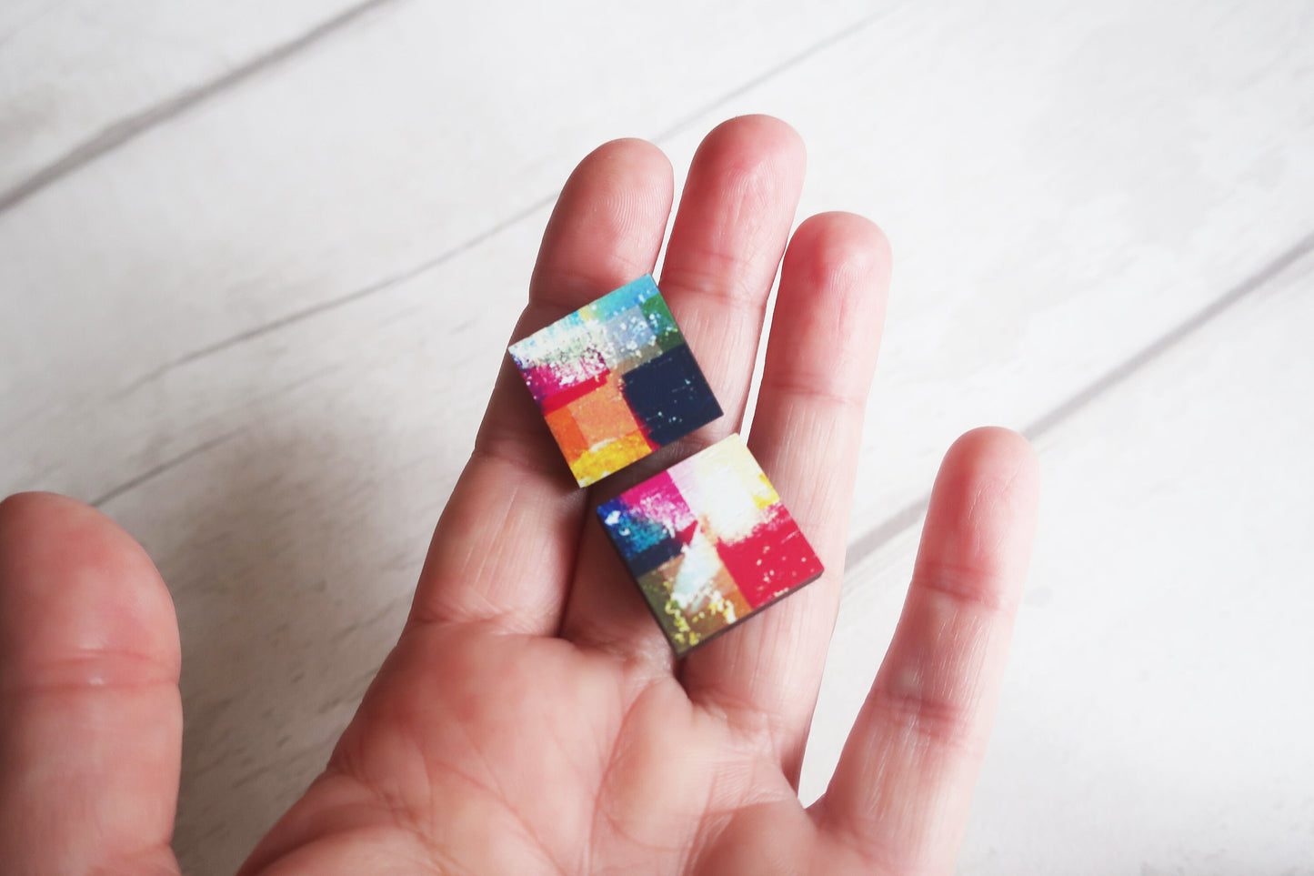 Colorful square earrings held in a hand on a light wooden surface