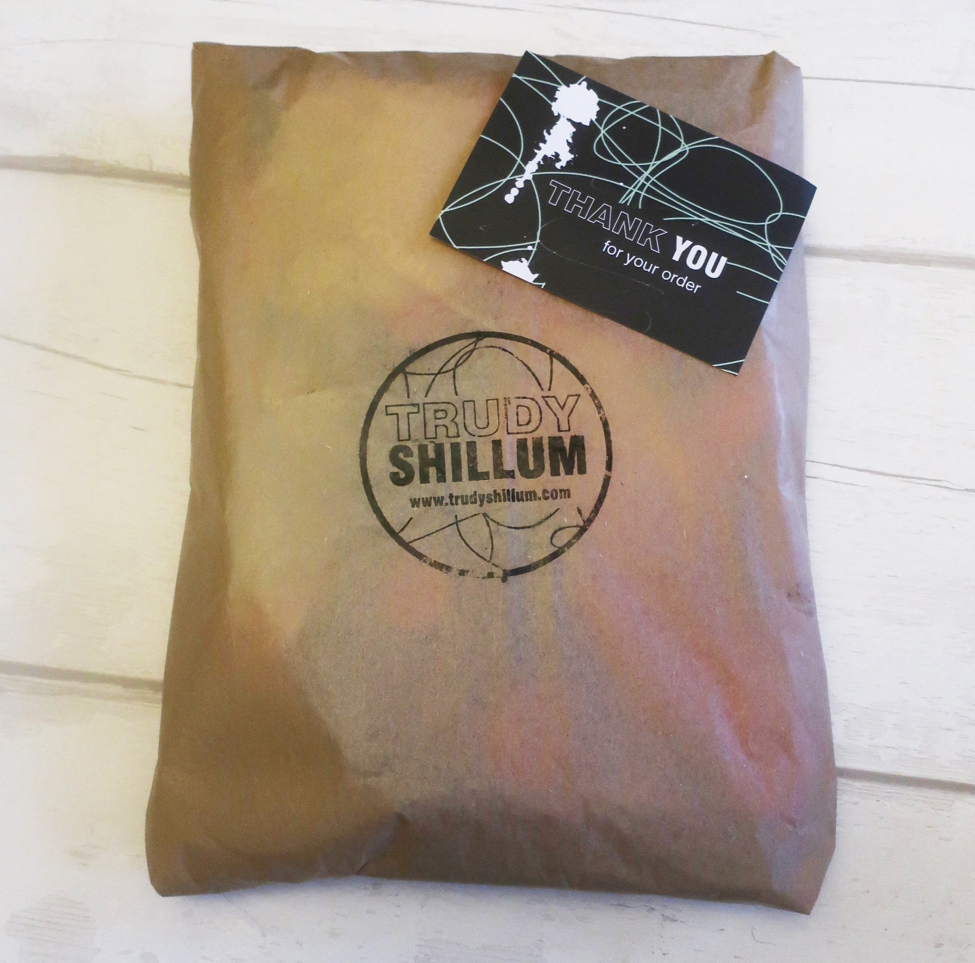Brown paper package with Trudy Shillum logo and a 'Thank You' card on a white wooden surface.