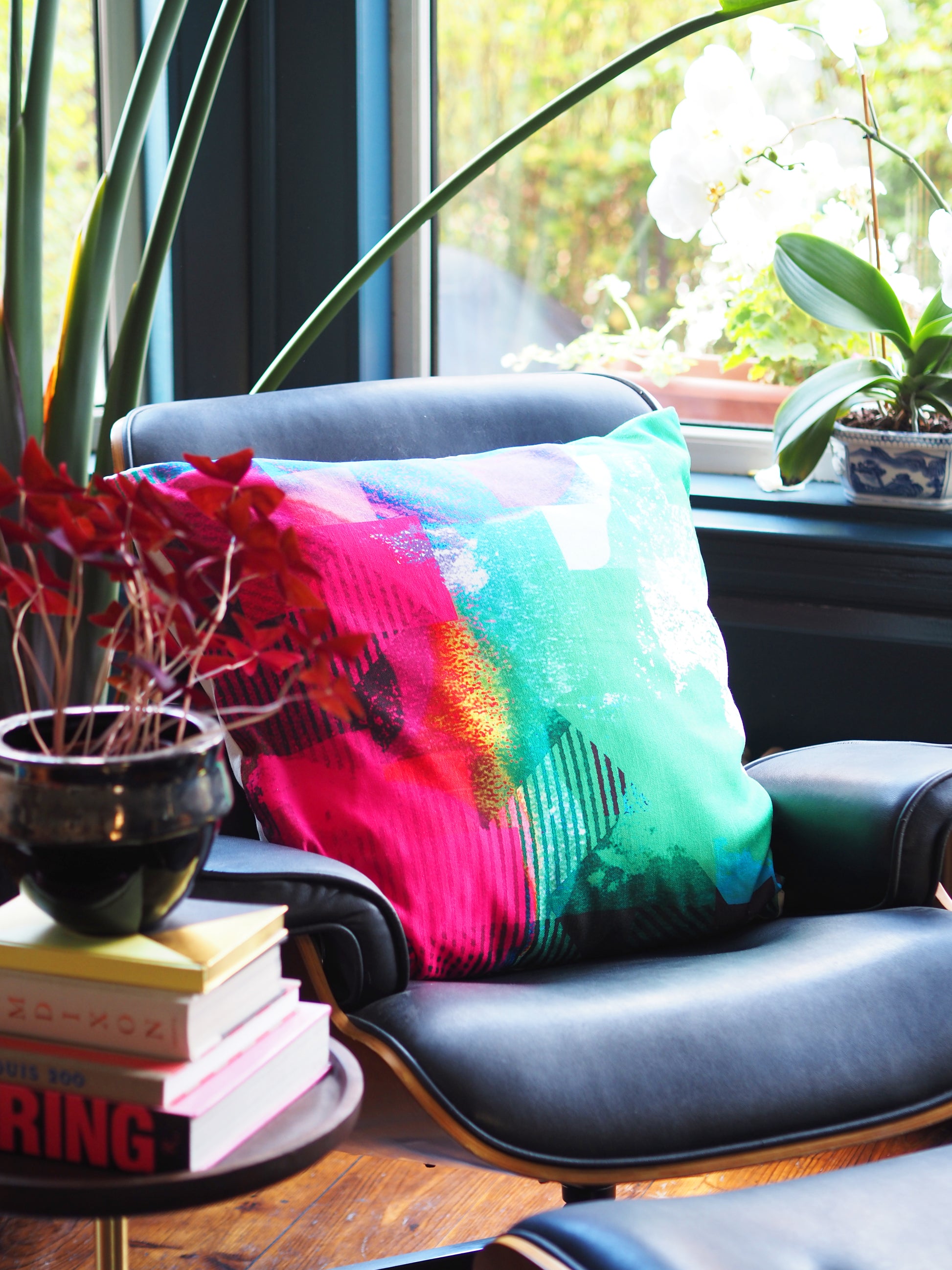Colorful pillow on a chair with books and plants in a room.