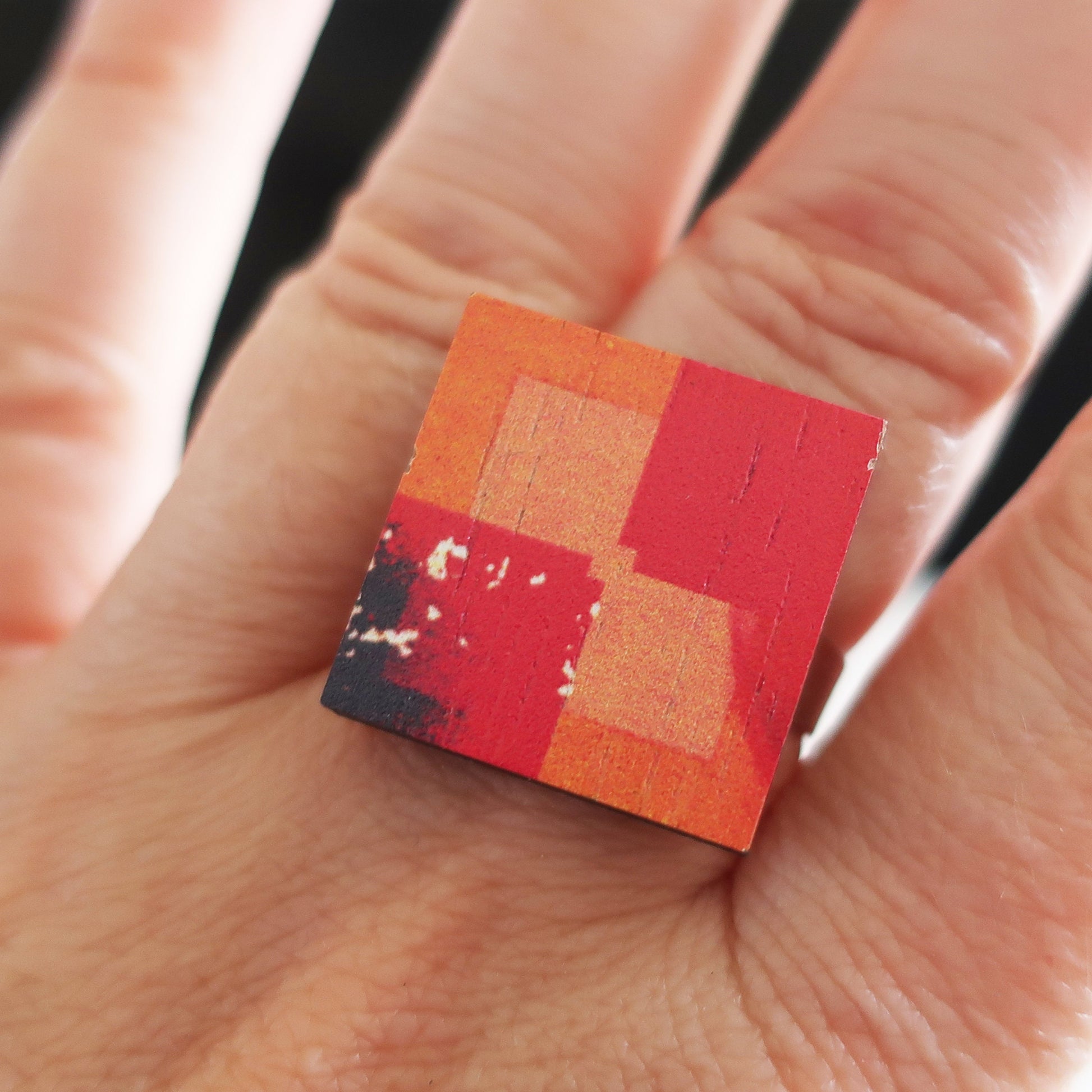 Small square wooden ring with red and orange checkered pattern held between fingers
