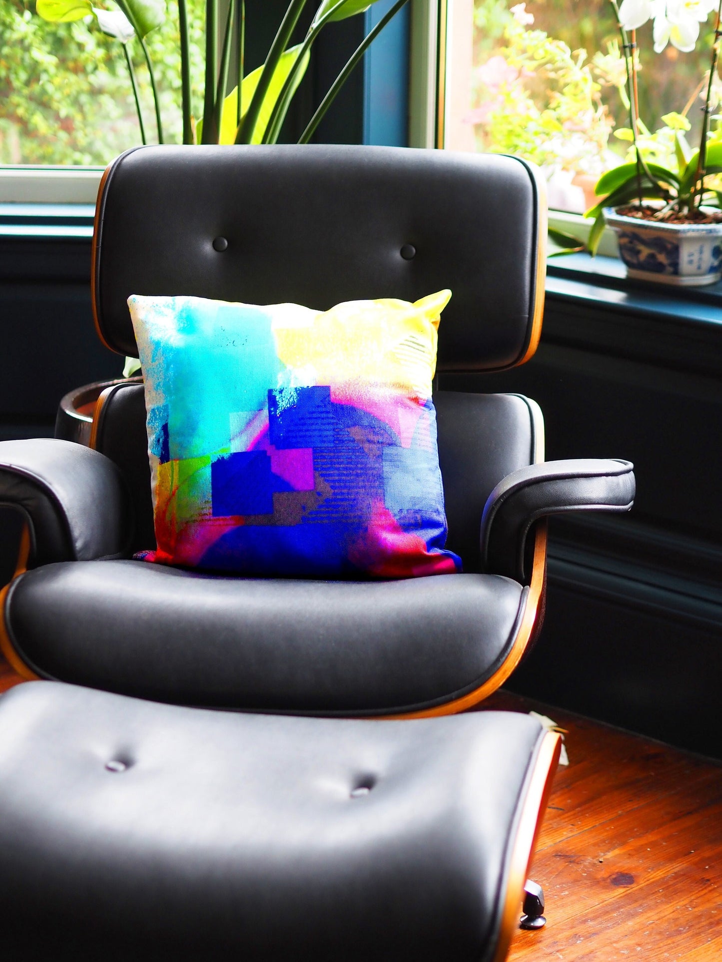 Colorful pillow on a black leather chair with a window and plants in the background