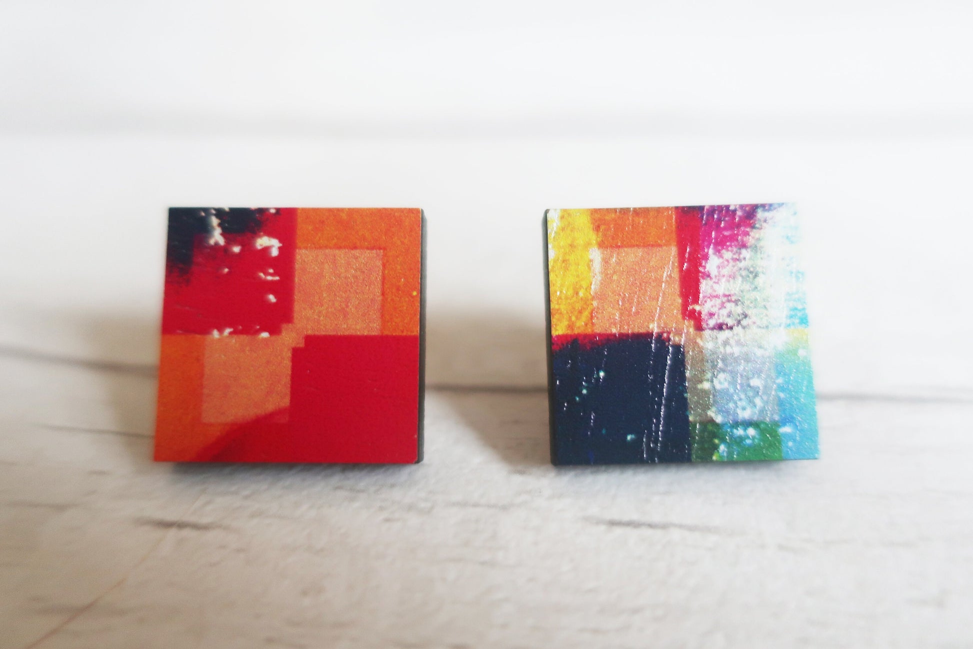 Two square earrings with colorful abstract designs on a light wooden surface.
