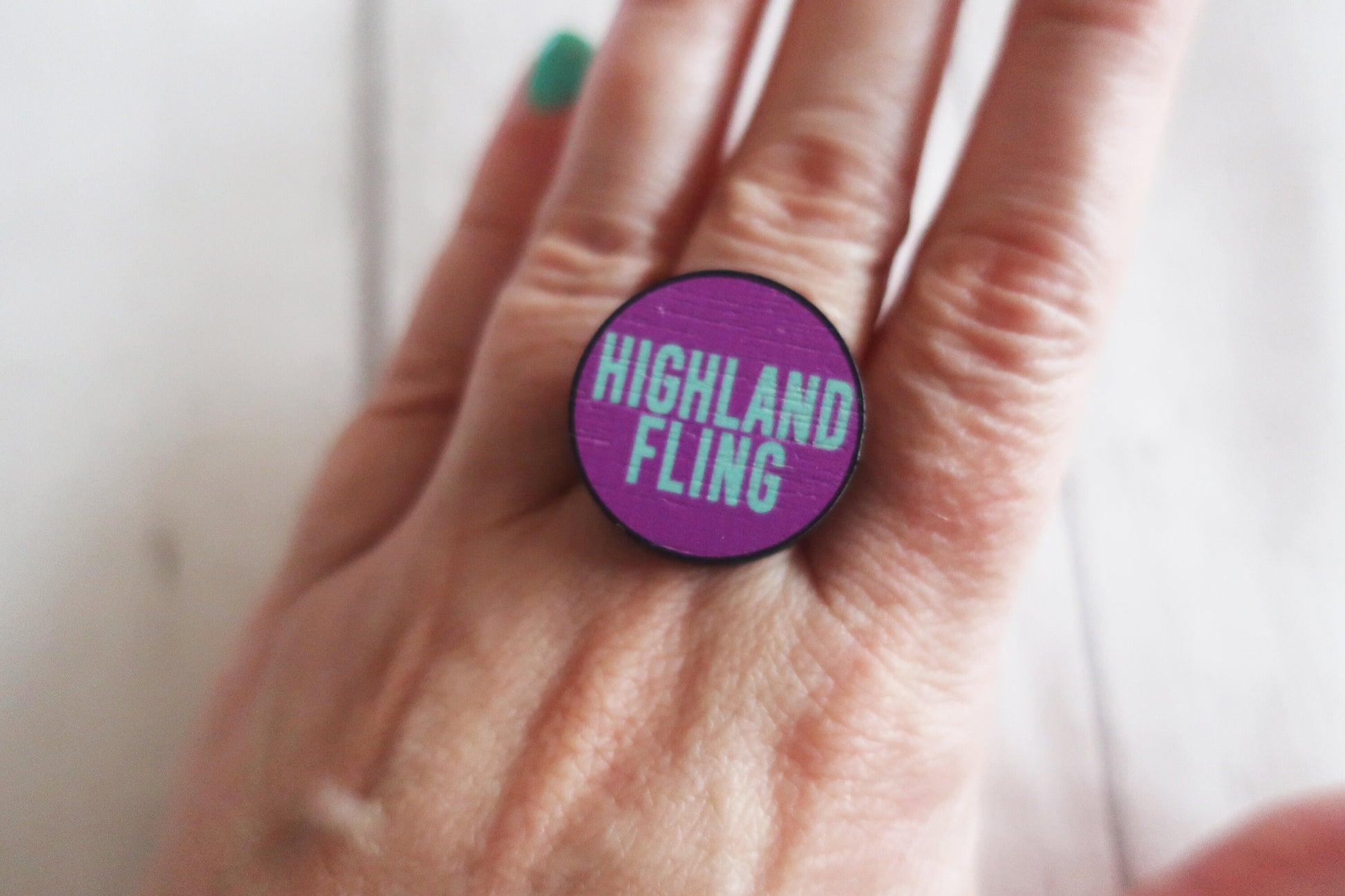 Hand wearing a purple ring with 'Highland Fling' text against a neutral background