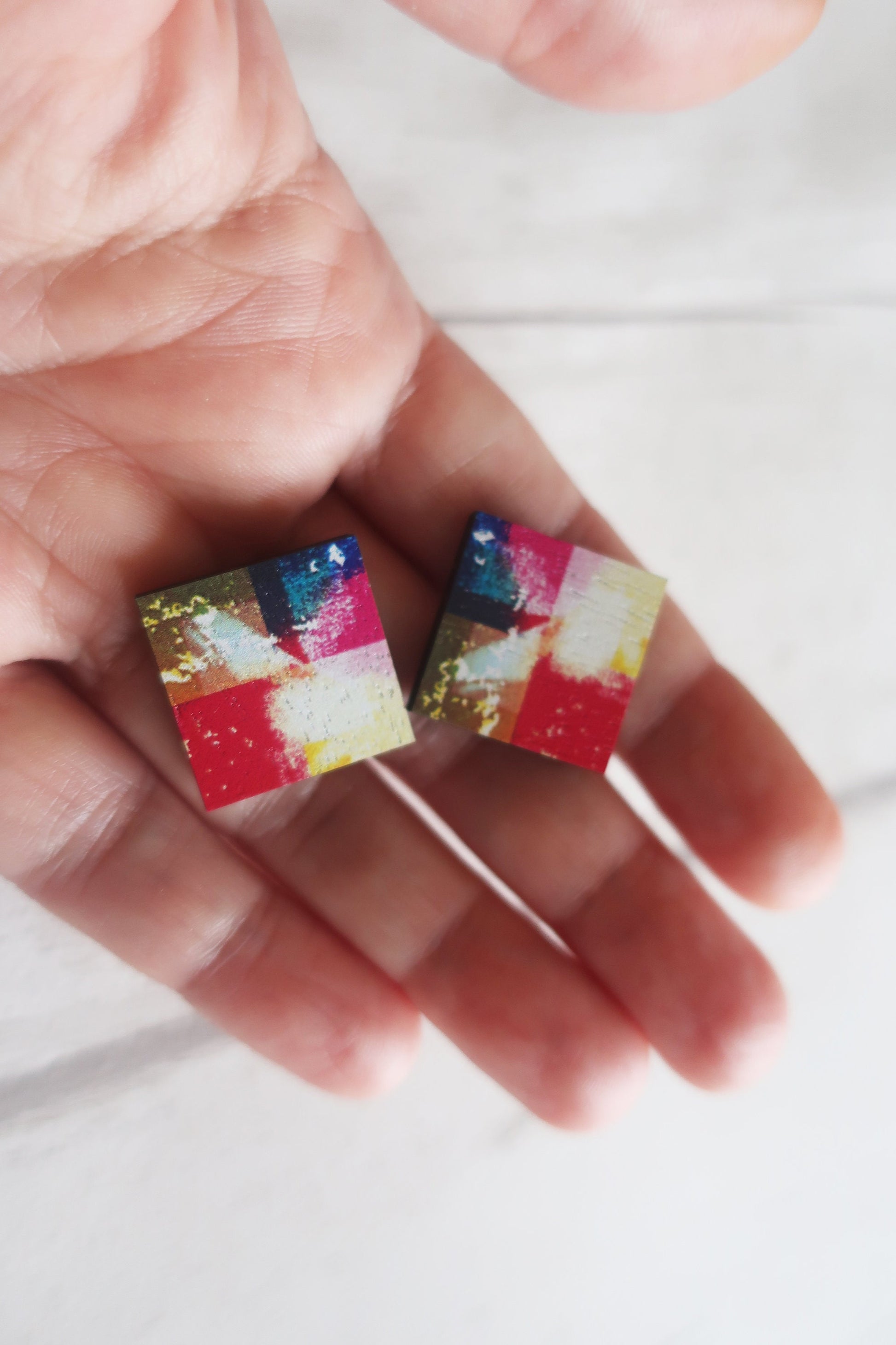 Colorful square earrings held between fingers on a white background