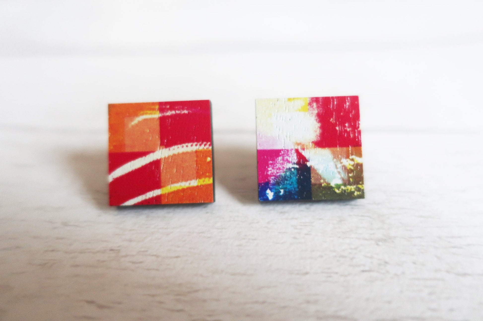 Two colorful square earrings on a light wooden surface.