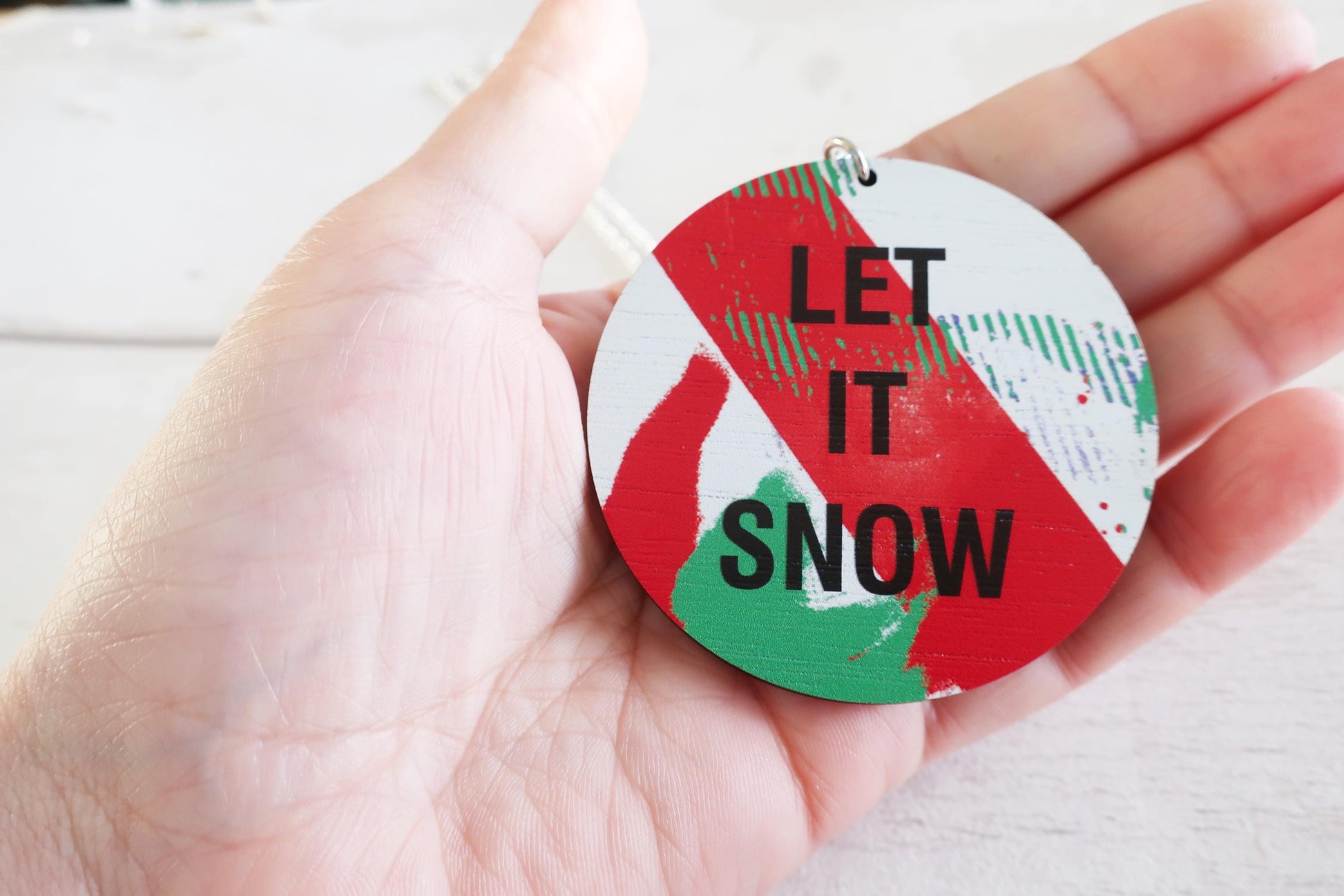 Hand holding a round decorative necklace  with 'LET IT SNOW' text on a white background