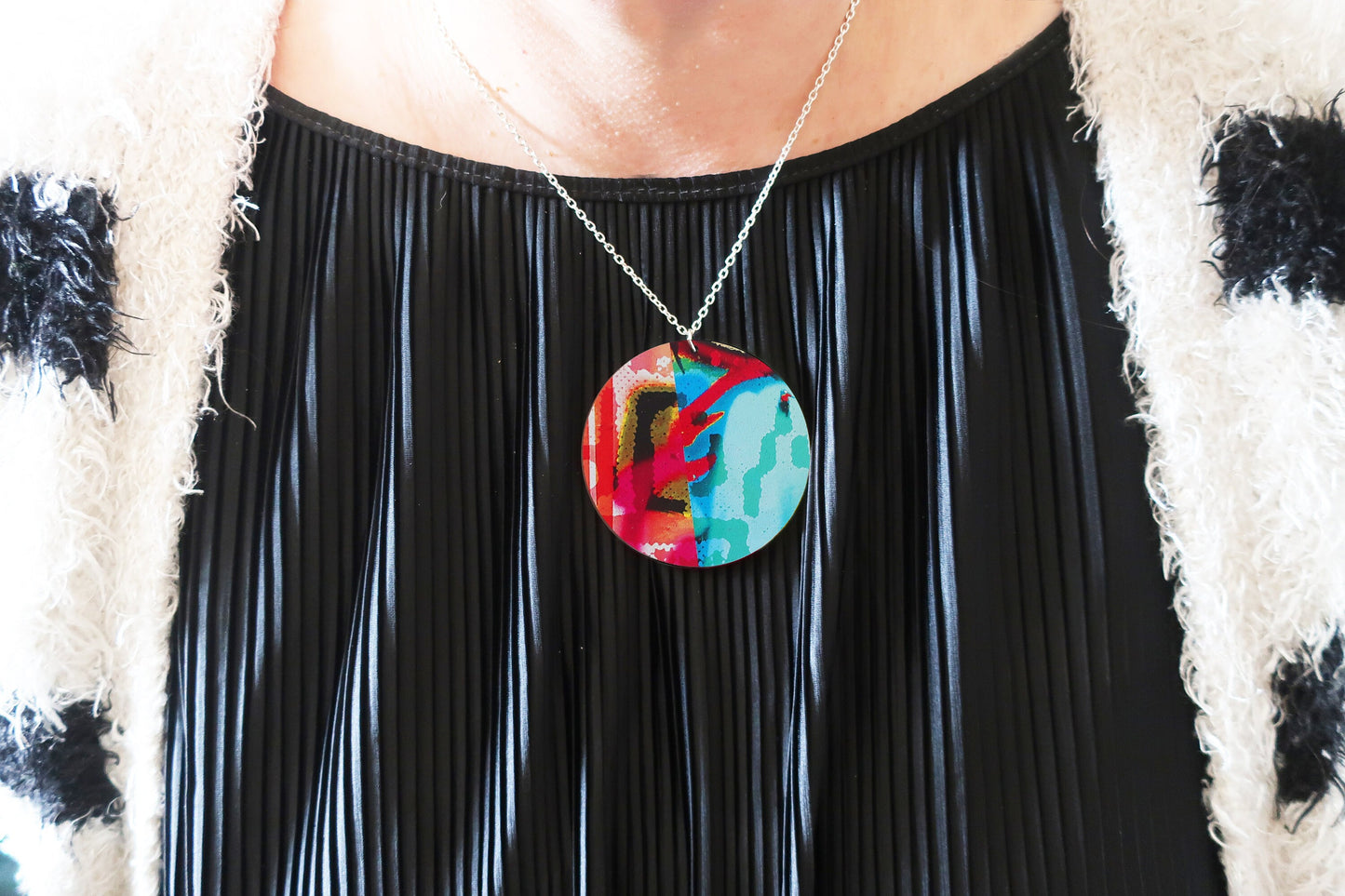 Person wearing a colorful pendant necklace with a black pleated dress and patterned sweater.