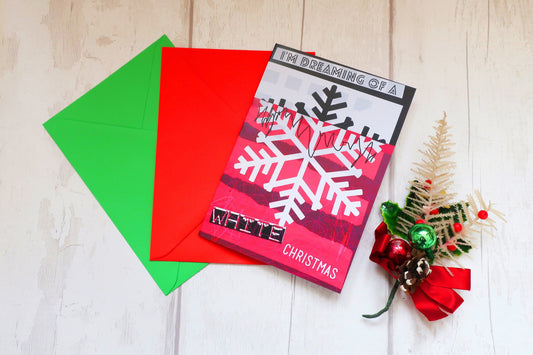Christmas card with snowflake design and red and green envelopes on a light wooden surface.