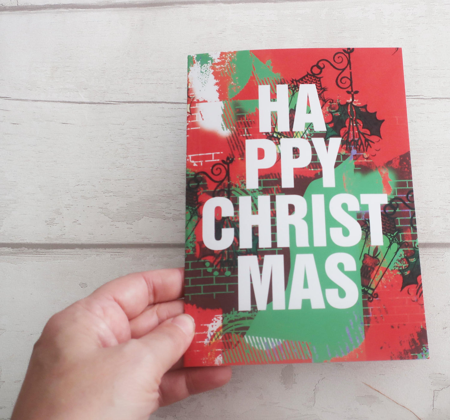 Hand holding a colorful Christmas card with 'Happy Christmas' text on a wooden surface