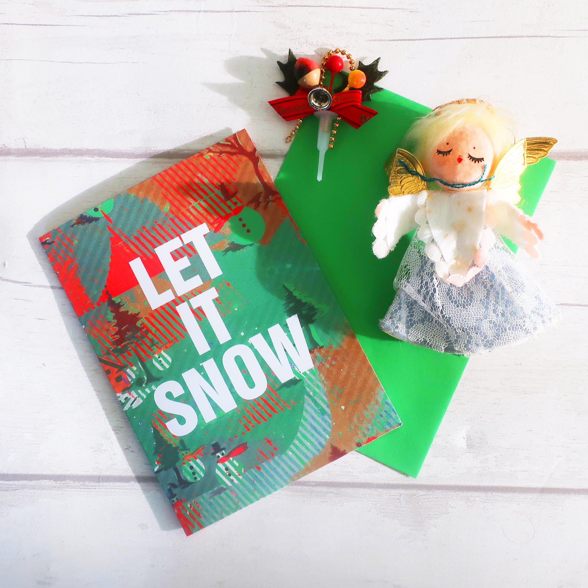 Christmas-themed card with 'LET IT SNOW' text and a small angel figurine on a white wooden surface.