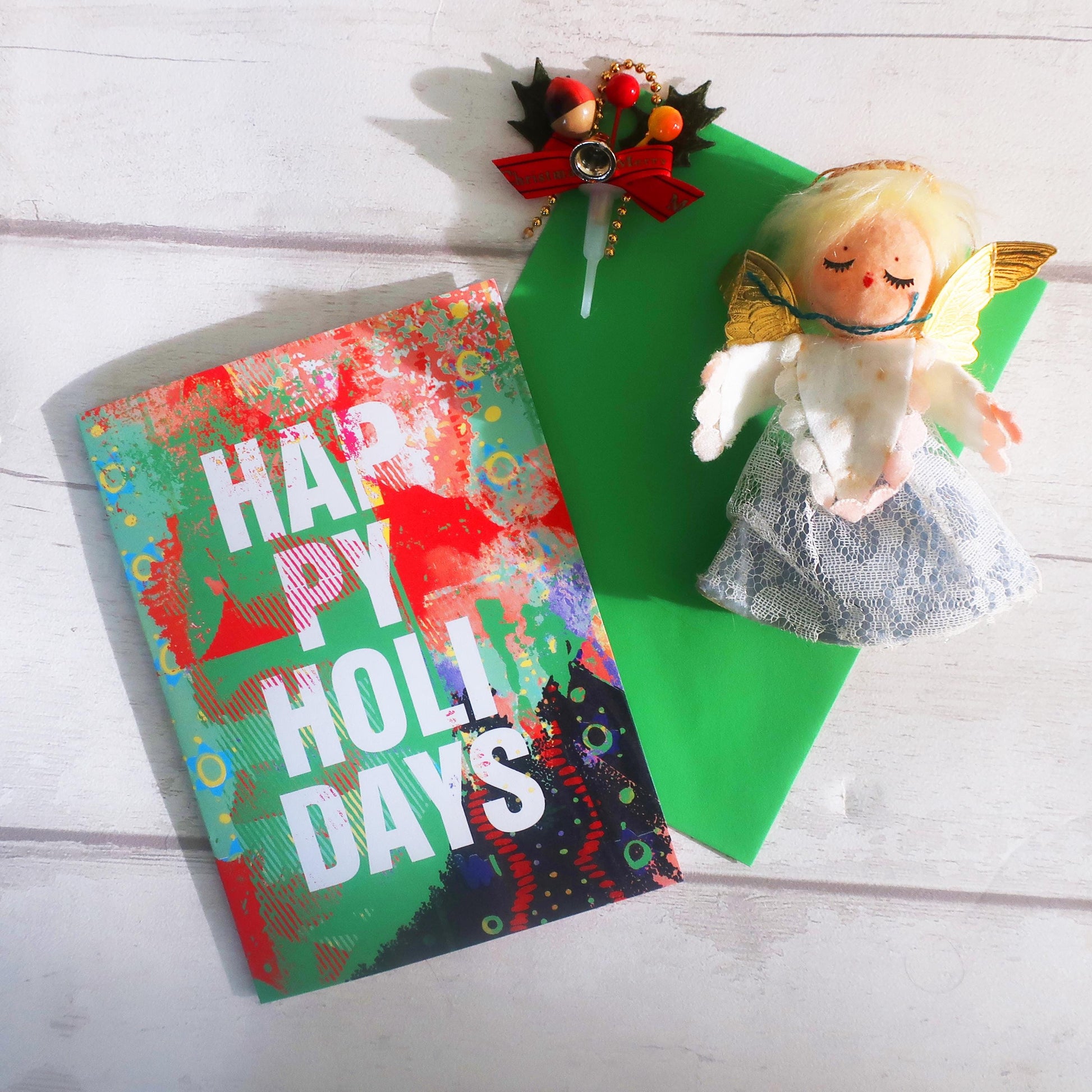 Colorful 'Happy Holidays' card with a decorative brooch and small angel figurine on a wooden surface.