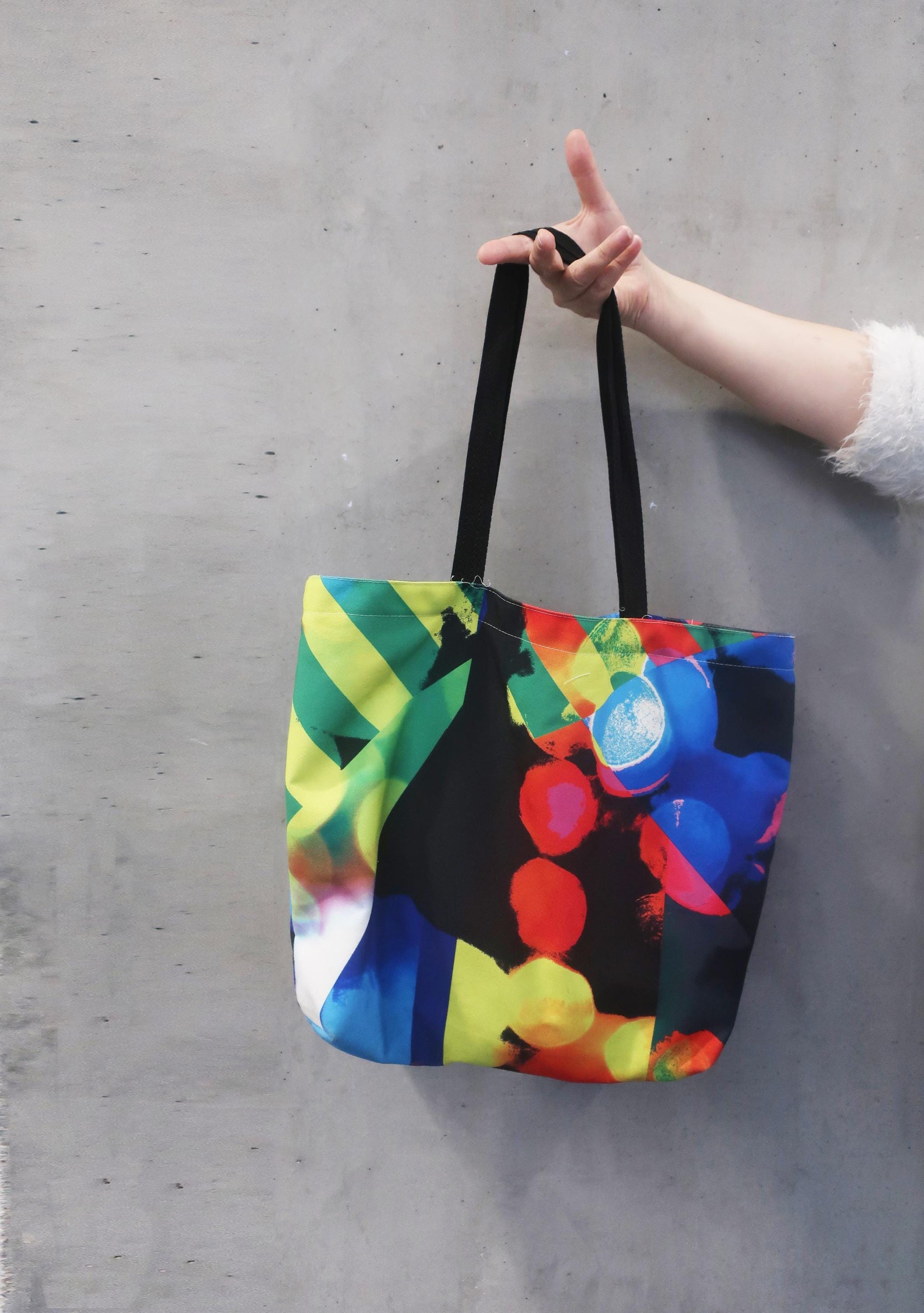 Colorful tote bag with abstract design held by a person on a gray concrete floor.