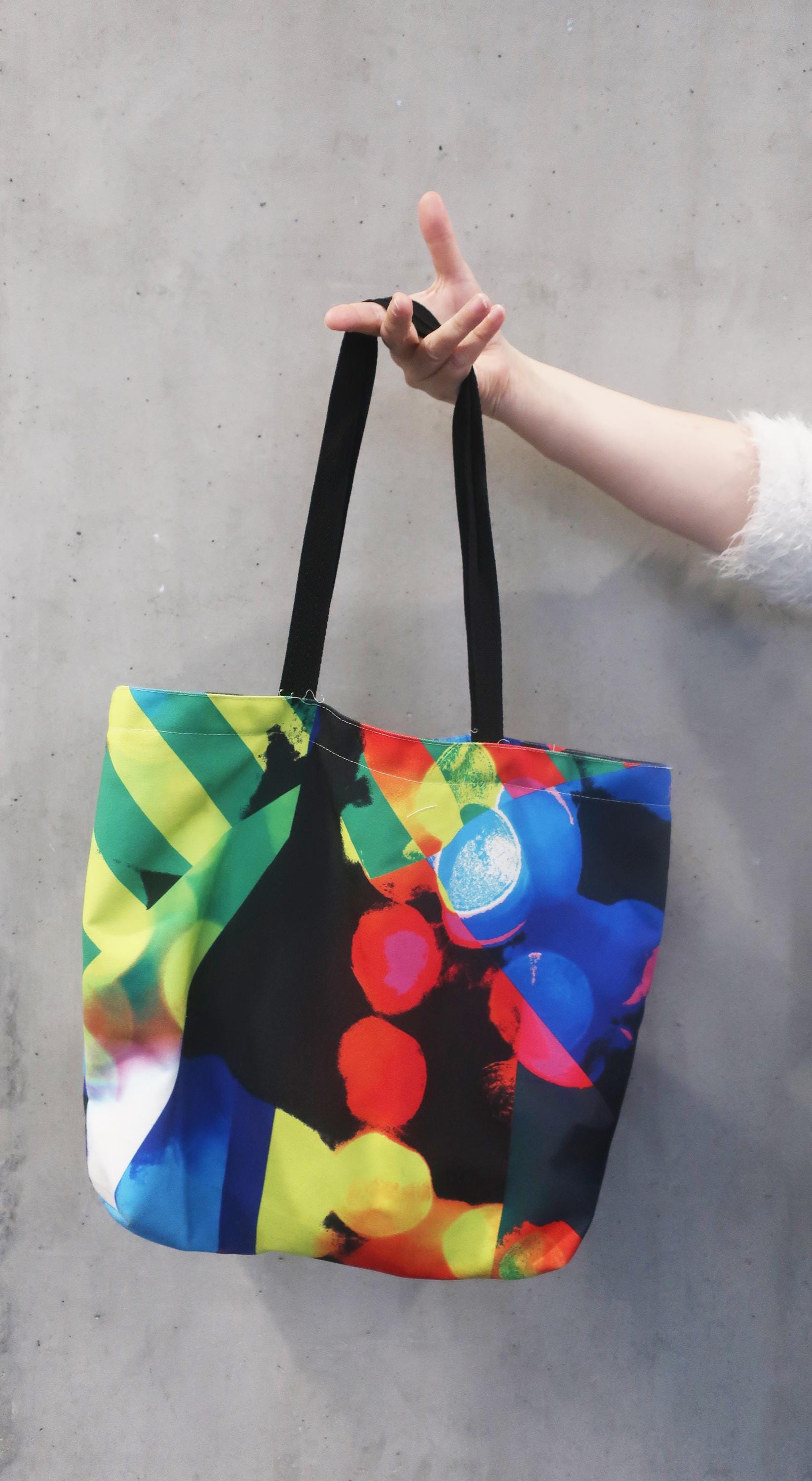 Colorful tote bag with black handles held by a person against a gray background
