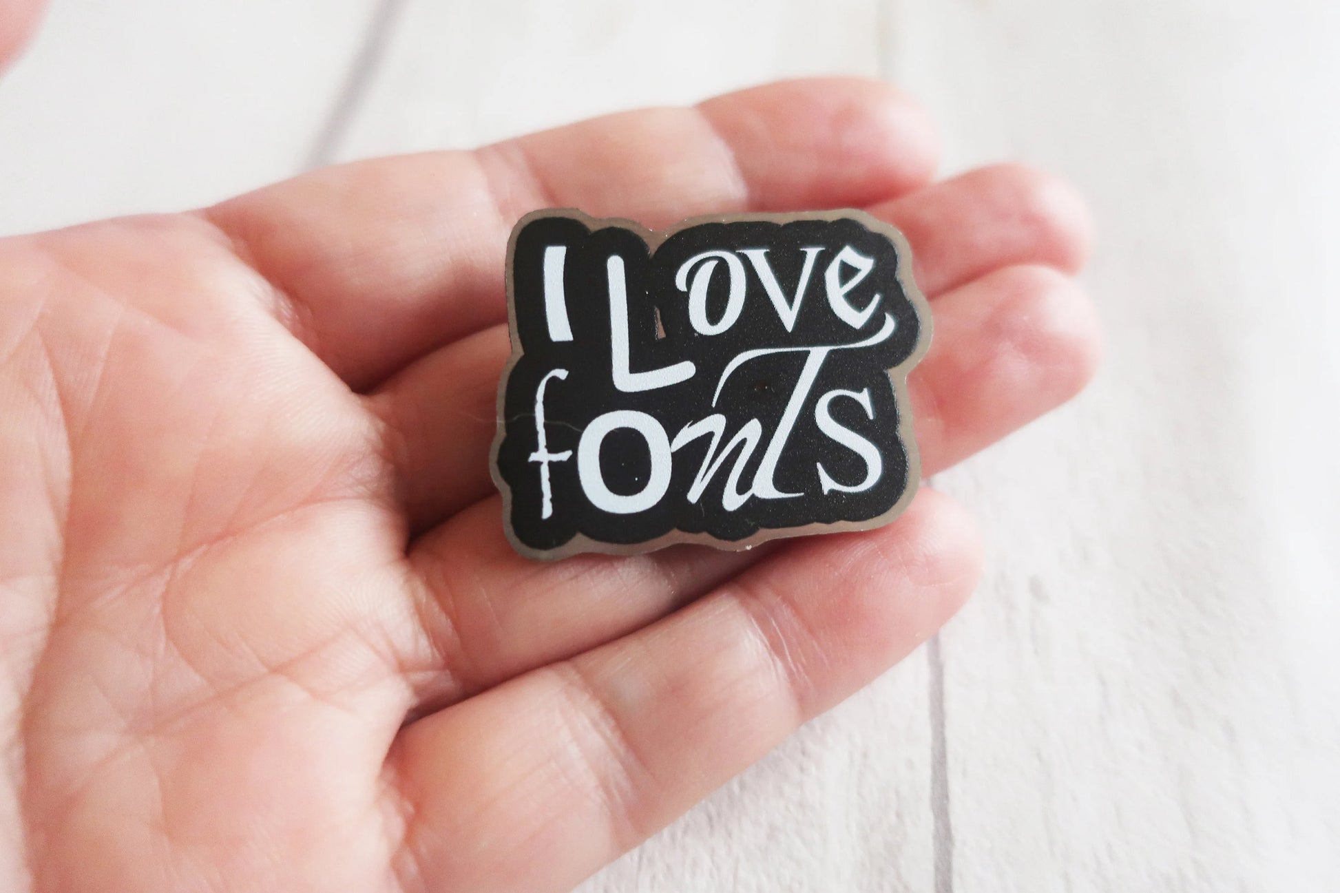 Pin with 'I Love Fonts' text held in a hand against a white background