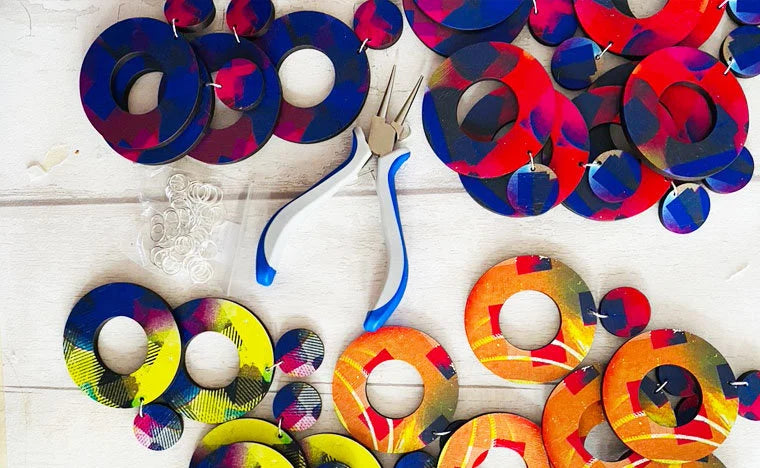 Colorful circular earrings being made on a white wooden surface with a pair of jewellery pliers.